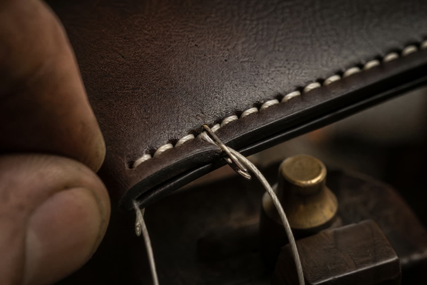 Macro of hand saddle stitching along a burnished wallet edge, thread visibly crossing within each hole