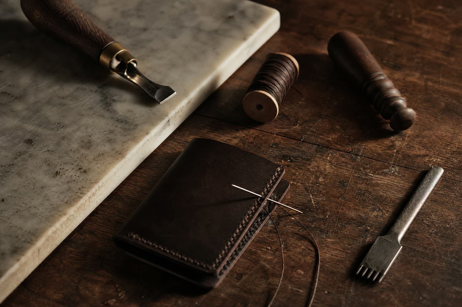 A leather workshop bench from above with a marble slab, edge beveller, wooden slicker, waxed thread and a half-stitched wallet with two needles still in the seam