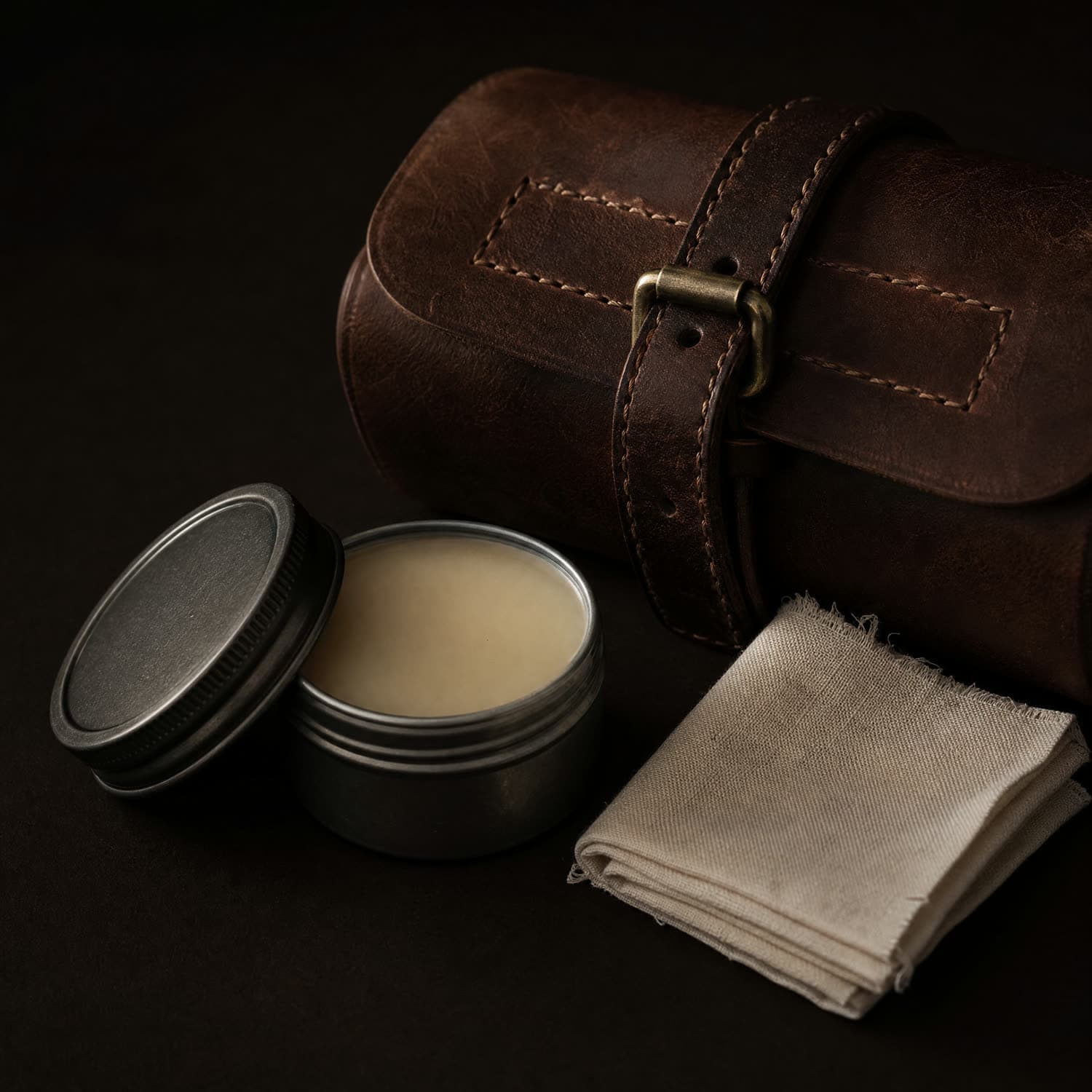 An open steel tin of pale neutral leather balm beside a folded undyed cotton cloth